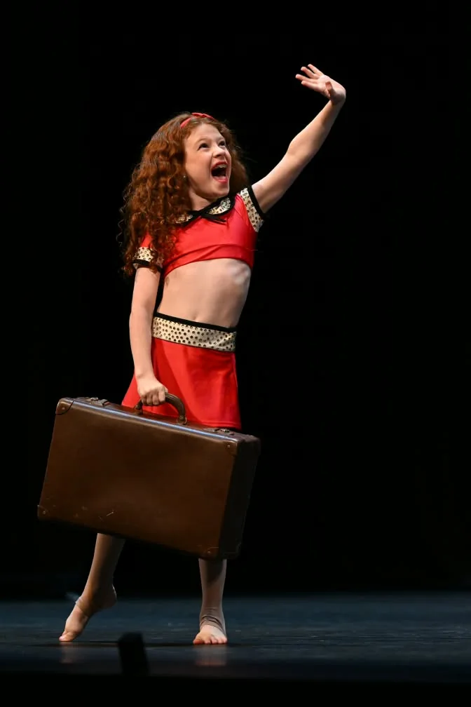 Young JMDS soloist in red, waving and carrying a suitcase