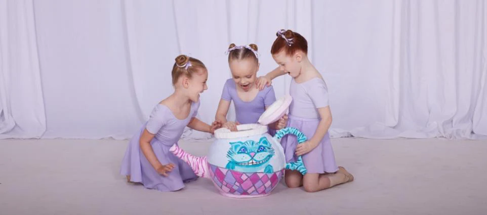 Three Tiny Tots in lilac leotards and skirts peeking into a painted Cheshire cat teapot