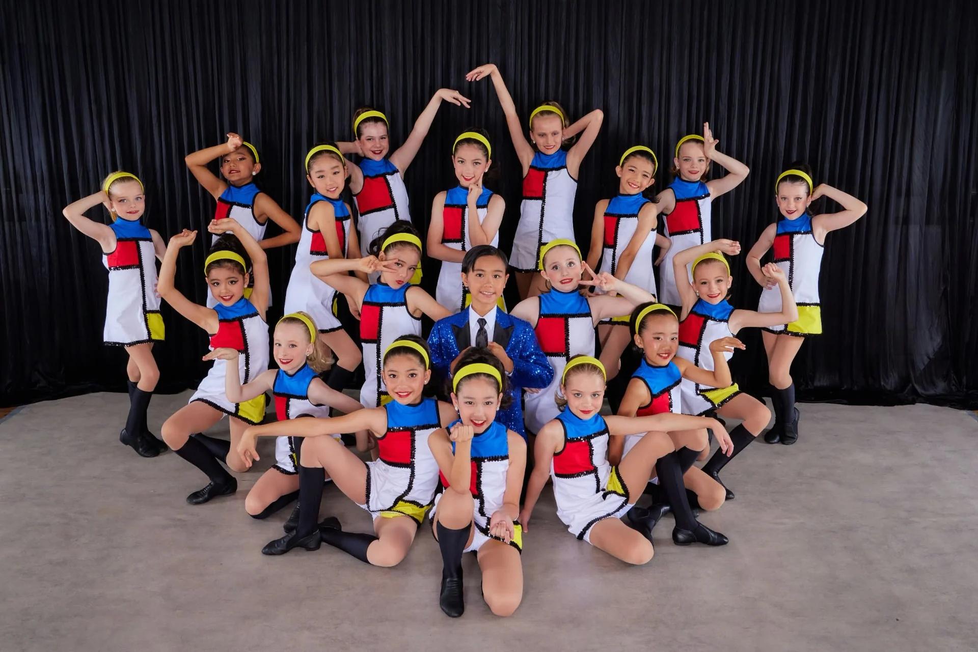 JMDS troupe in colour-block mod dresses and yellow headbands posing around a dancer in a sequinned blue jacket